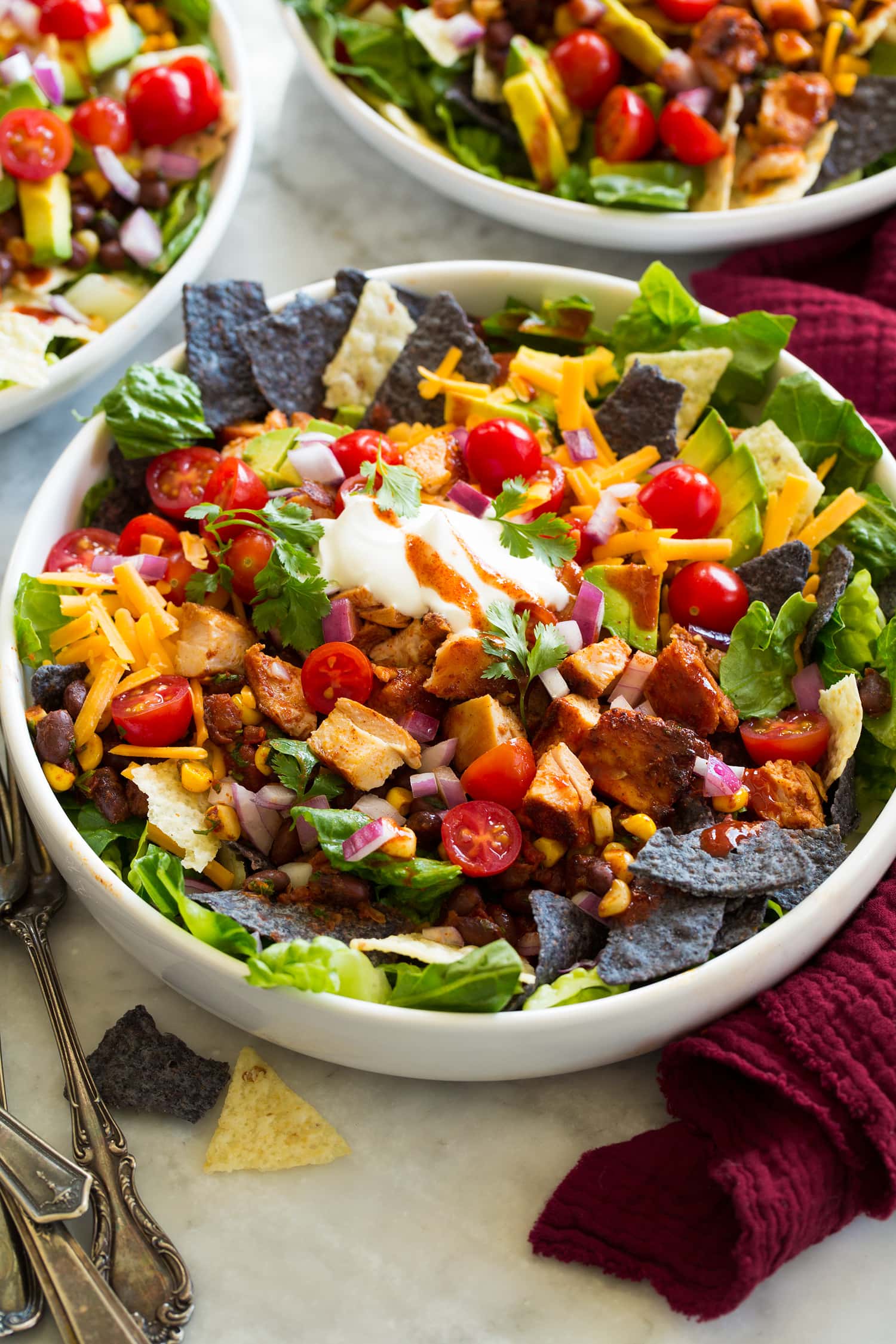 Chicken taco salad shown with blue and yellow tortilla chips, corn, avocado, tomatoes, lettuce, chicken, cilantro, cheese and sour cream.